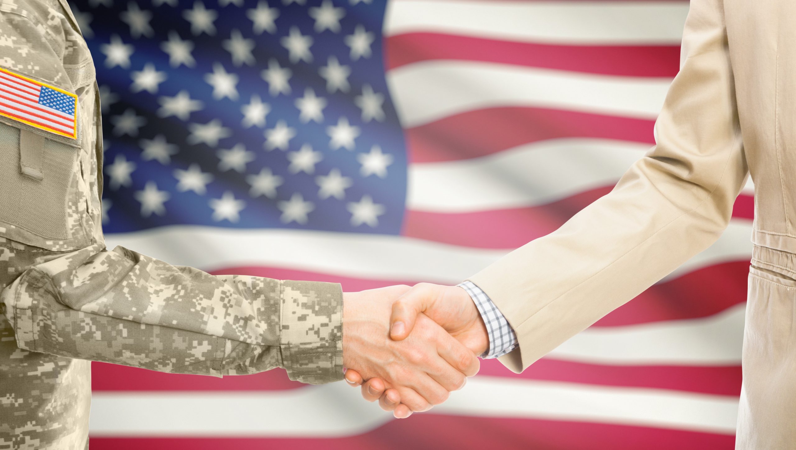 USA military man in uniform and civil man in suit shaking hands with adequate national flag on background - United States of America American soldier in uniform and civil man in suit shaking hands with adequate national flag on background - United States of America